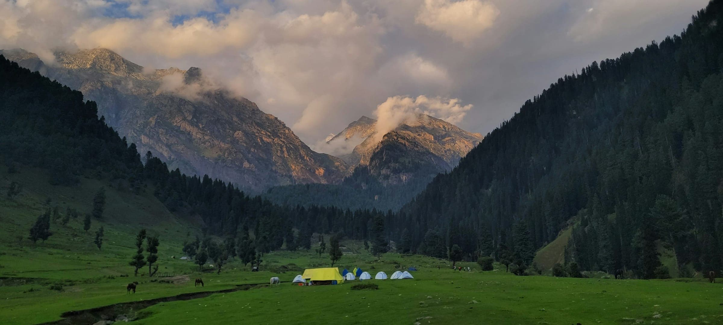 Golden evening light breaking through clouds over a green Kashmir valley, with a meadow camp below