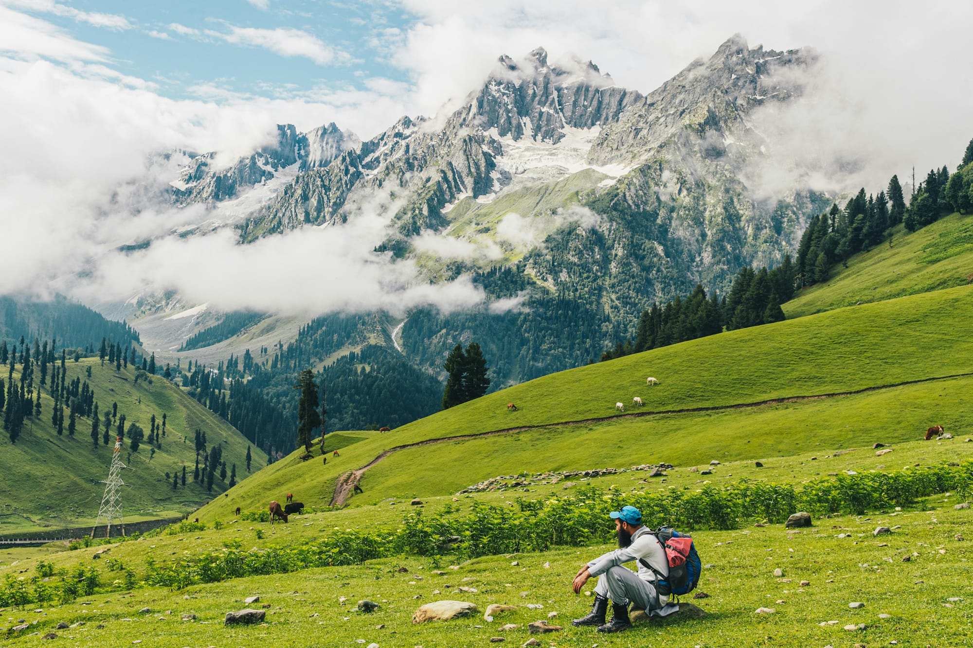 A trekker resting on a green slope beneath jagged, cloud-wrapped Kashmir peaks
