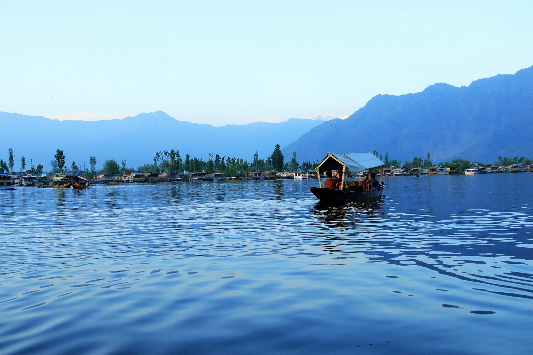 A shikara crossing the still, blue waters of Dal Lake at dusk with houseboats along the shore